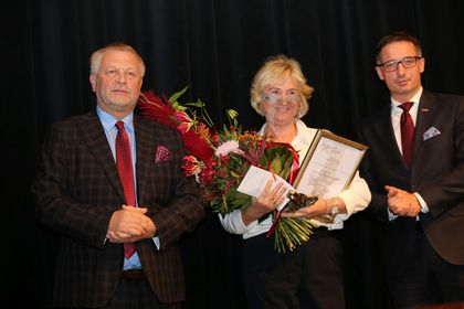 Na zdjęciu trzy osoby, dwaj mężczyźni w ciemnych garniturach, w środku ubrana w jasne kolory kobieta trzymająca w rękach dyplom, statuetkę i kwiaty. 