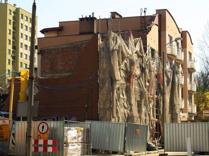 Remont kamienicy. Fasada budynku zasłonięta płachtami. Przed budynkiem blaszany płot. 