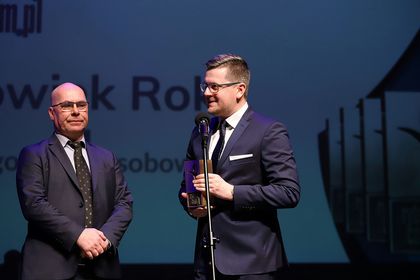 Dwóch mężczyzn w ciemnych garniturach i okularach. Jeden trzyma statuetkę. 