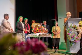 Grupa osób stoi na scenie. W tle widać bukiety kwiatów.