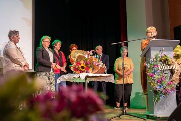 Grupa osób stoi na scenie. W tle widać bukiety kwiatów.