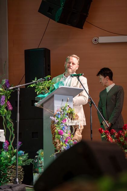 Na zdjęciu widać dwie osoby na scenie. Na pierwszym planie jest mężczyzna, który mówi do mikrofonu.