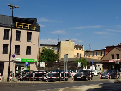 Wybrukowany placyk w centrum miasta. Na nim zaparkowane samochody. Wokół kamienice. 