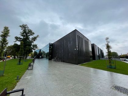 Nowoczesny budynek i prowadząca do niego szeroka aleja. W tle zachmurzone niebo i zieleń. 