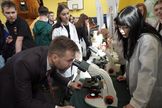 Grupa osób w sali gimnastycznej. Na pierwszym planie dwójka osób. Mężczyzna patrzy w mikroskop. 