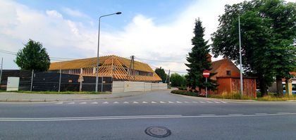 Droga. Po lewej stronie za płotem budynek w trakcie remontu dachu. Po prawej stronie zieleń i ceglane zabudowania. 