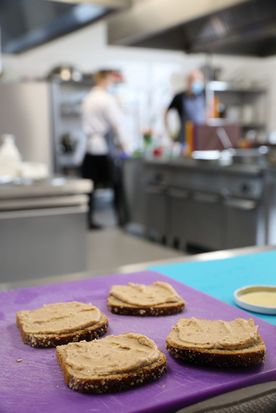 Kanapki na fioletowej serwetce na kuchennym blacie, w tle osoby pracujące w kuchni. 