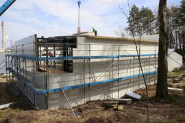 Budynek kontenerowy otoczony rusztowaniem w trakcie rozbudowy. W tle niebo, zalew i kominy elektrowni.