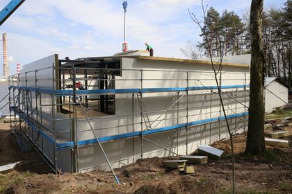 Budynek kontenerowy otoczony rusztowaniem w trakcie rozbudowy. W tle niebo, zalew i kominy elektrowni.