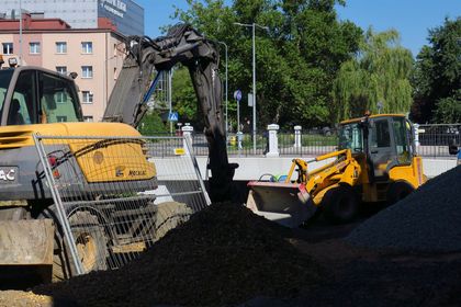 Roboty budowlane na drodze wzdłuż rzeki. W tle most, wokół drzewa i zabudowa miejska. 