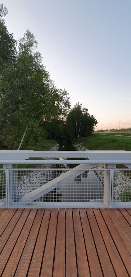 Balustrada mostu na rzece. Po obu stronach zieleń. Po prawej stronie ekrany akustyczne i barierki przy drodze szybkiego ruchu. 
