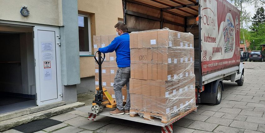 Maseczki fot 1 Mężczyzna w niebieskiej bluzie rozładowuje paczki z auta dostawczego przy pomocy wózka widłowego.
