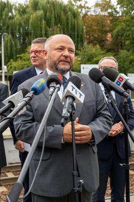 Zdjęcie przedstawia mężczyznę mówiącego do mikrofonu. W tle widać dwóch mężczyzn.