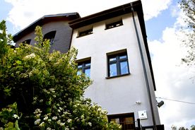 Jasny budynek na tle nieba, wokół zieleń. 