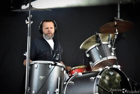 Mężczyzna w ciemnym ubraniu i słuchawkach prze perkusji w trakcie koncertu. 