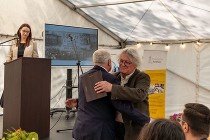 Na zdjęciu widać dwóch mężczyzn na pierwszym planie, którzy wykonują uścisk w ramach oficjalnego spotkania. W tle widać kobietę.