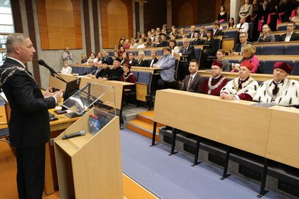 Mężczyzna przemawiający do mikrofonu w auli. W tle siedzący ludzie. 