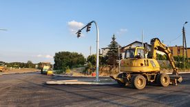 Droga w trakcie remontu, świeżo położony asfalt. Wzdłuż drogi domy, zieleń.