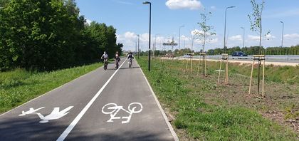 Trzy rowerzystki w maseczkach na drodze rowerowej. Po obu stronach zieleń. W dali, po prawej stronie, ekrany akustyczne i barierki przy drodze szybkiego ruchu. 