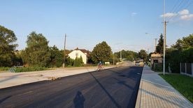 Droga w trakcie remontu, świeżo położony asfalt. Wzdłuż drogi domy, zieleń.