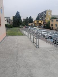 OsrodekJPII remont 17 Podjazd dla osób niepełnosprawnych. W tle zabudowania i szare niebo, po prawej stronie zaparkowane samochody, na wprost trawnik.