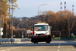 Ulice miasta, na nich pługopiaskarka i pracownicy służb komunalnych z szuflami do odśnieżania.