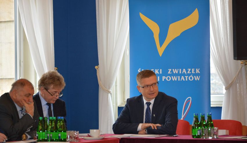 Trzy osoby siedzą przy stole narad. W tle logo Śląskiego Związku Gmin i Powiatów