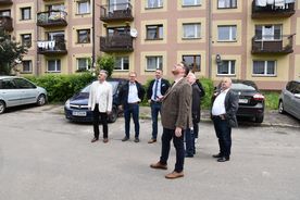 Grupa mężczyzn na osiedlowym parkingu patrzy na coś do góry. W tle samochody i zabudowania.