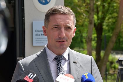 Mężczyzna w jasnym garniturze - prezydent Rybnika - na tle budynku i zieleni. Przed nim mikrofony.