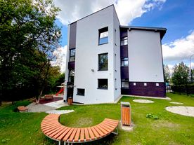 Jasny dwupiętrowy budynek na tle błękitnego nieba, wokół zieleń. 