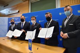 Pięciu mężczyzn w ciemnych garniturach i maseczkach na twarzy w trakcie konferencji prasowej prezentuje podpisany list intencyjny. W tle banery reklamowe miasta Rybnika i subregionu zachodniego. 