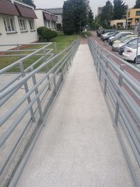 OsrodekJPII remont 20 Podjazd dla osób niepełnosprawnych. W tle zabudowania i szare niebo, po prawej stronie zaparkowane samochody, po lewej trawnik.