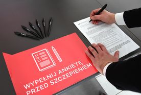 Leżąca na stole karta z wzorem deklaracji do wypełnienia. Z boku dłonie trzymające długopis. Obok kilka długopisów i czerwona