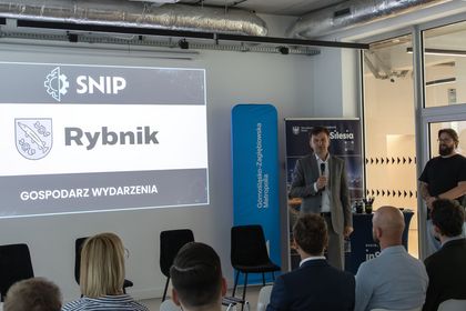 Zdjęcie przedstawia mężczyznę z mikrofonem. W tle ekran i siedzący ludzie. 