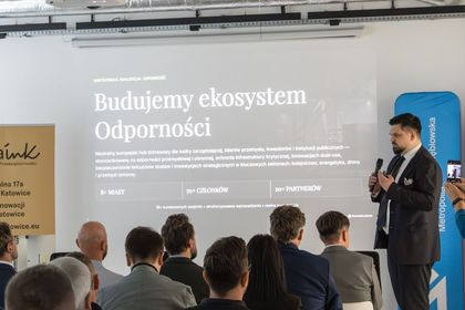 Zdjęcie przedstawia mężczyznę z mikrofonem. W tle ekran i siedzący ludzie. 