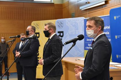 Pięciu mężczyzn w ciemnych garniturach i maseczkach na twarzy w trakcie konferencji prasowej. W tle banery reklamowe miasta Rybnika i subregionu zachodniego. 