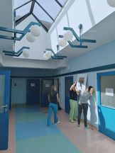 OsrodekJPII remont 7 Trzy kobiety wieszają obraz w antyramie na ścianie. Przestronny, jasny korytarz, po obu stornach otwarte drzwi w kolorze niebieskim, podwieszany sufit ze świetlikami okiennymi.