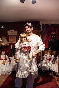 Zdjęcie przedstawia mężczyznę trzymającego w rękach straszną lalkę. W tle widać inne straszne lalki.