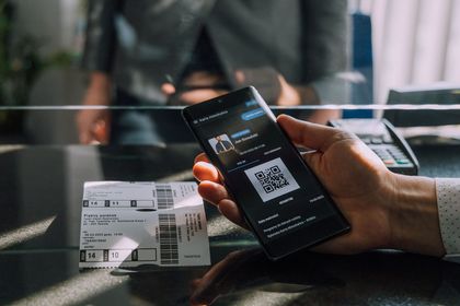 Smartfon z wyświetlonym kodem QR na tle okienka kasowego, widoczna też ręka obsługująca urządzenie 