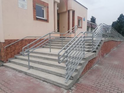 OsrodekJPII remont 16 Wyremontowane schody z podłożem lastrikowym i niklowanymi poręczami prowadzące do niskiego jasnego budynku. Obok chodnik z kostki betonowej.