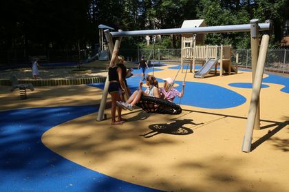 Plenerowy plac zabaw, w otoczeniu zieleni parkowej. Piaskownica, drwniana jeżdżalnia, huśtawki, bawiące się dzieci. 