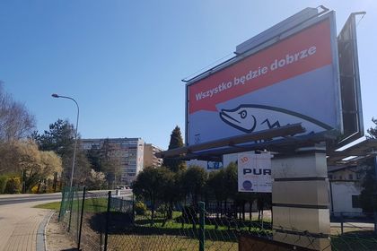 Zebrzydowicka 3 Po prawej stronie billboard na tle błękitnego nieba, po lewej ulica, chodnik i blok w tle.