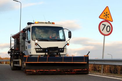 Ulice miasta, na nich pługopiaskarka i pracownicy służb komunalnych z szuflami do odśnieżania.
