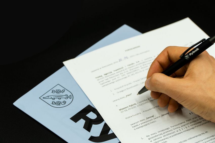 Dłoń z długopisem nad fragmentem porozumienia, o którym mowa w tekście na tle teczki z napisem Rybnik i herbem miasta na ciemnym tle. 