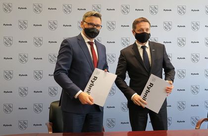 Prezydent Rybnika Piotr Kuczera i wójt Pilchowic Maciej Gogulla podpisują umowę przy stole konferenycyjnym. W tle ścianka reklamowa z herbem Rybnika