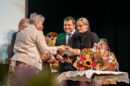 Na zdjęciu widać grupę osób. W tle widać bukiety kwiatów.