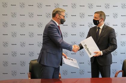 Prezydent Rybnika Piotr Kuczera i wójt Pilchowic Maciej Gogulla podpisują umowę przy stole konferenycyjnym. W tle ścianka reklamowa z herbem Rybnika