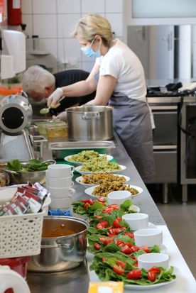 Różnokolorowe potrawy na talerzach na metalowym kuchennym blacie, w tle dwie pracujące w kuchni osoby