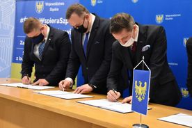 Mężczyźni w ciemnych garniturach i maseczkach na twarzy podpisują list intencyjny. W tle banery reklamowe miasta Rybnika i subregionu zachodniego. 