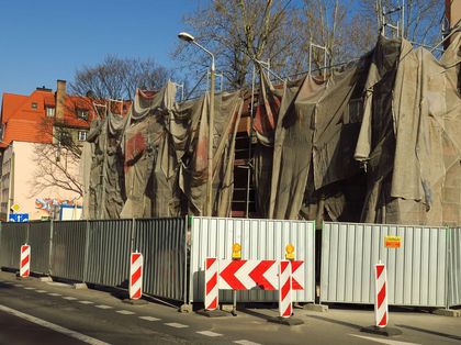 Remont kamienicy. Fasada budynku zasłonięta płachtami. Przed budynkiem blaszany płot. 